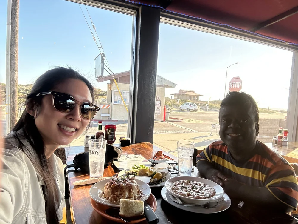 Anita Wing Lee and Tim Muttoo sharing a meal together at a seaside restaurant, laughing